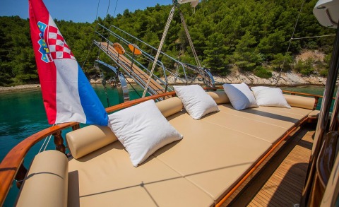 Sunlit aft deck with beige cushions and white pillows, wooden rails, Croatian flag, calm blue-green water and green forest in the background