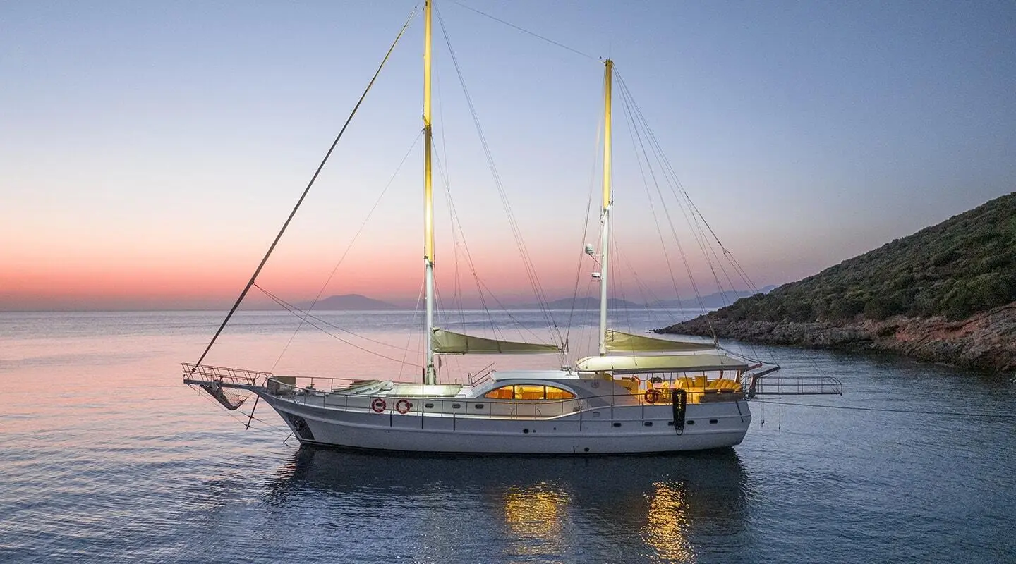 White two-masted sailing yacht anchored near rocky coast at sunset
