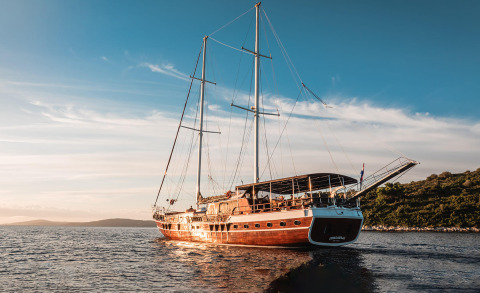 Wooden sailing yacht with people on the aft deck under a canopy, on calm sea at sunset