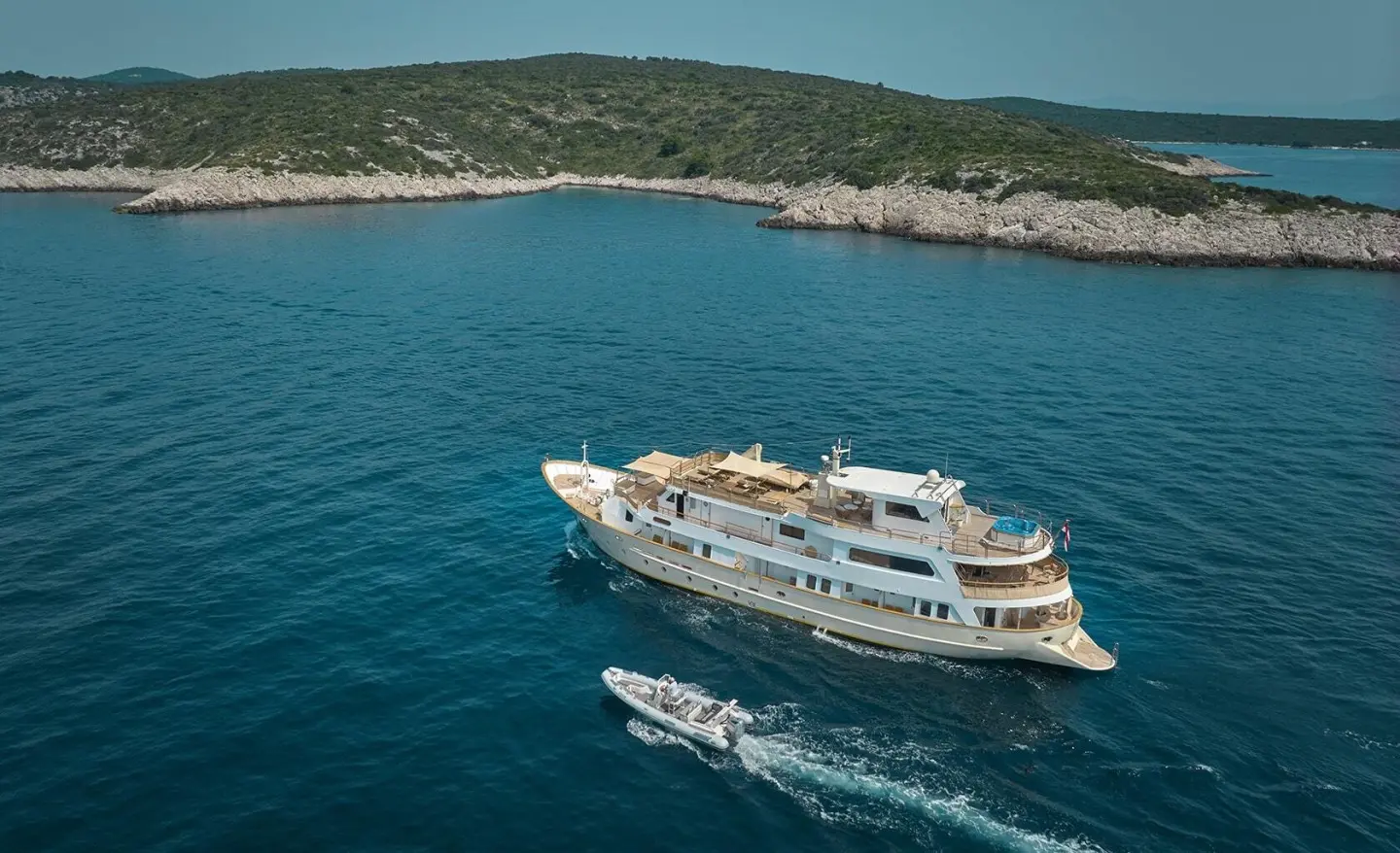 A white luxury yacht named La Perla sails near a rocky coastline on blue water