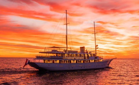 Luxurious sailing yacht Bellezza at sunset on the sea, with two masts, stern flag, and illuminated deck windows.