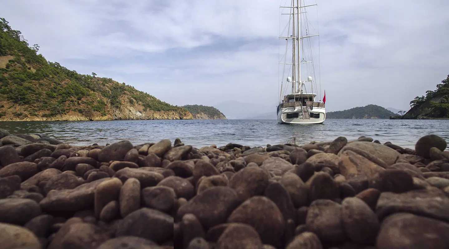 Kayhan 3 yacht anchored near rocky shore with a sailing hull and sunset sky