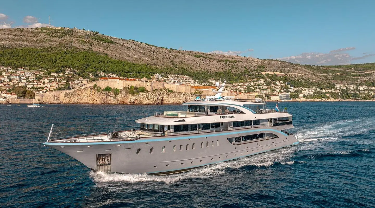 Luxury motor yacht Freedom gliding on blue water with rocky coastline behind