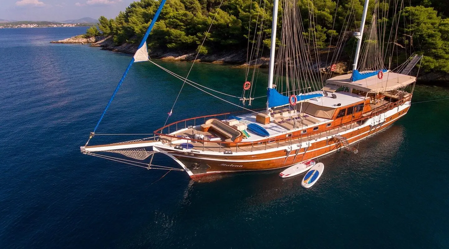 Malena yacht anchored in turquoise water at sunset with sails tied back and wooden hull