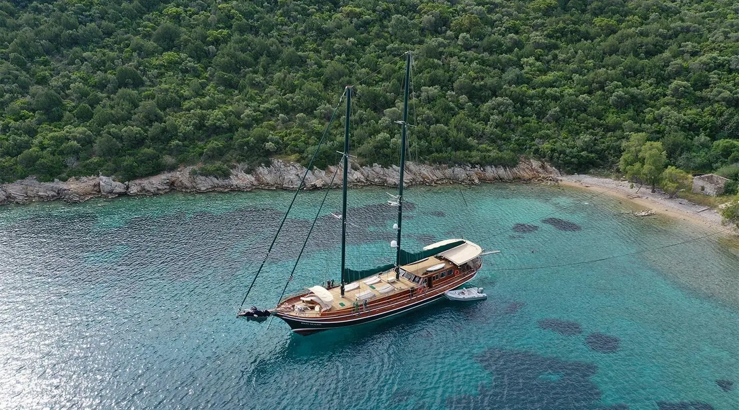 Gulet Ros Mare anchored in turquoise bay with forested hillside backdrop