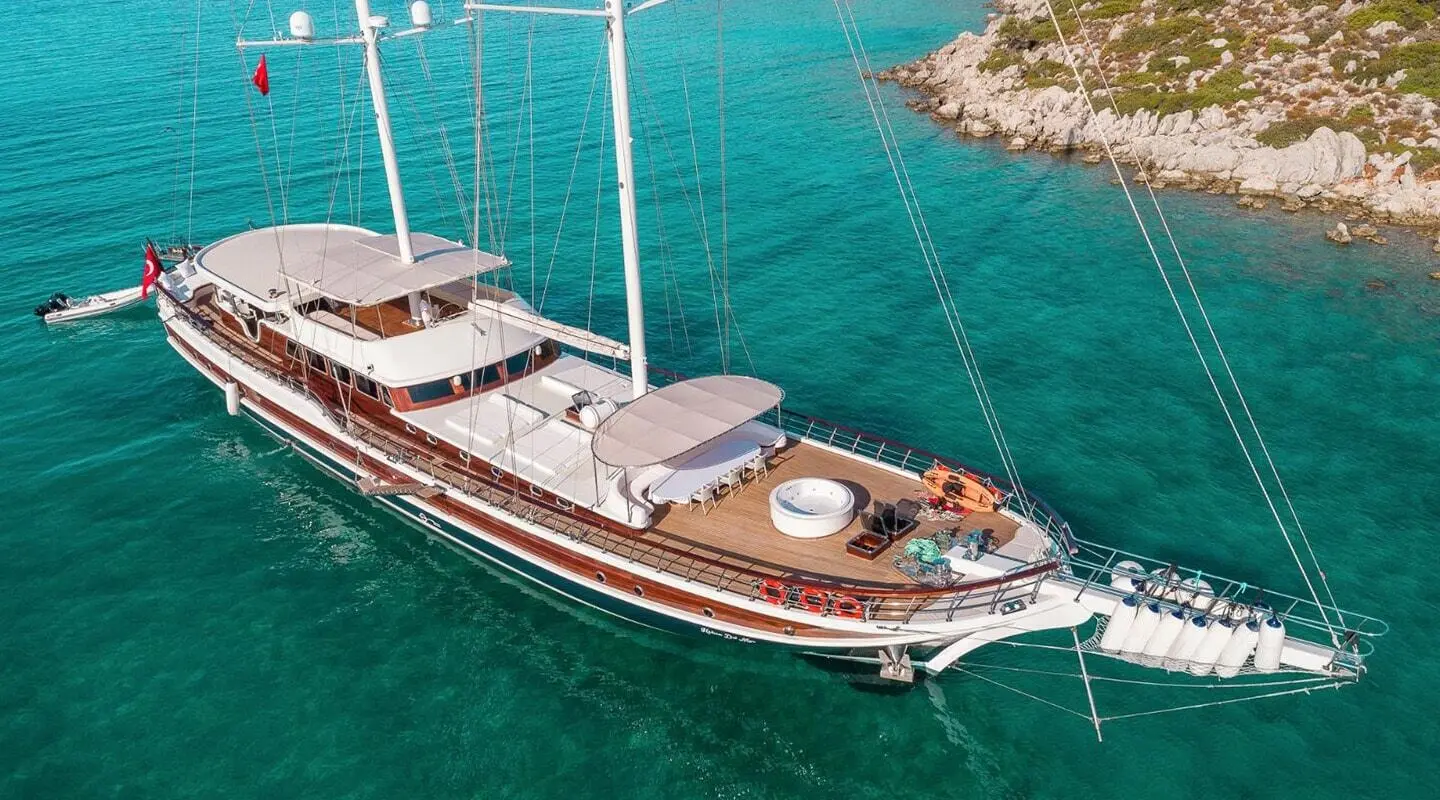 Large sailing yacht on turquoise water with deck and seating areas near rocky shore