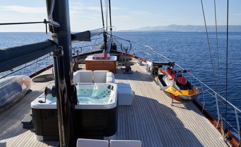 Sunlit open sea with a white seating area and a jacuzzi on the deck, a jet ski at the bow and distant coastline.