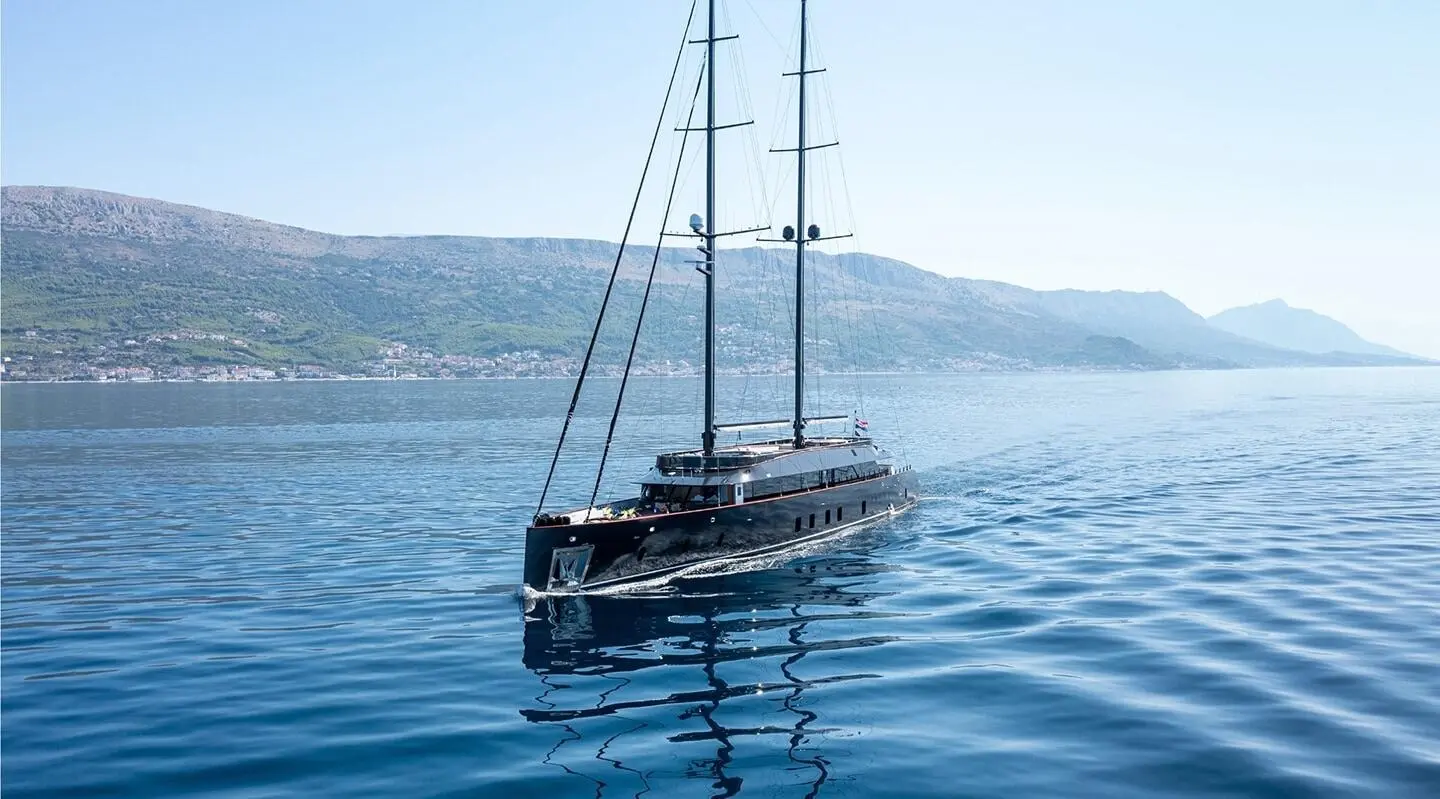 Yacht Scorpios cruising on calm blue water with sails lowered, coastal hills in background