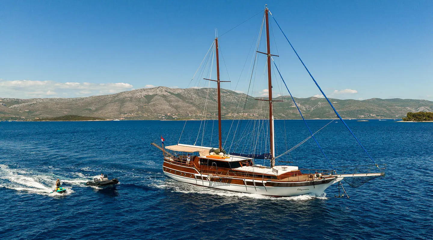 Twin-masted gulet named Eleganza anchored in calm blue sea against distant rocky coastline