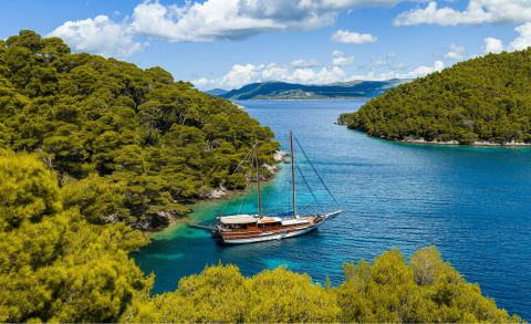 Wooden gulet yacht anchored in clear blue water beside green forested rocky coast