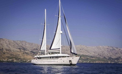 Omnia luxury sailing yacht anchored in calm blue waters with a rocky coastline in the background