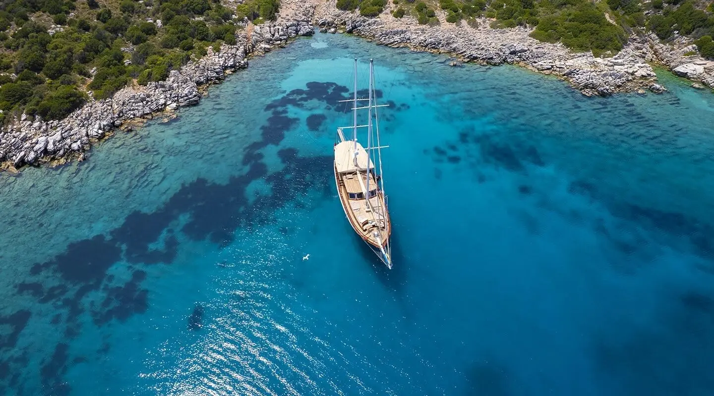 Wooden gulet yacht anchored in a clear blue bay with rocky shore nearby