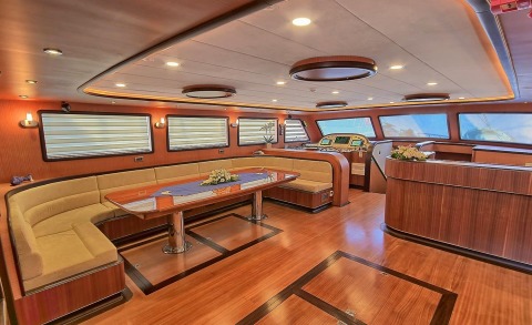Interior of a teak-furnished yacht saloon with a curved beige sofa and a wooden table