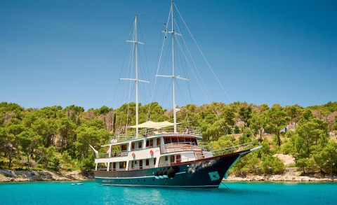 Sailing yacht with two masts anchored in turquoise water near green tree-covered shore