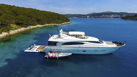 White motor yacht anchored in clear blue water near shore, people on upper deck