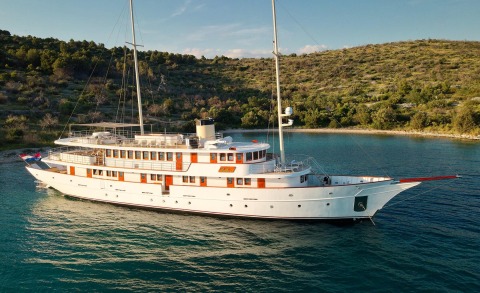 White luxury yacht Bellezza anchored near a rocky shoreline with windows along the side