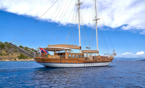 Wooden yacht sailing with a sunshade on the deck, with a ladder and railings, blue sea and distant land under a clear sky