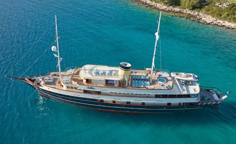 Large motor yacht with two decks anchored in clear turquoise water beside rocky shore
