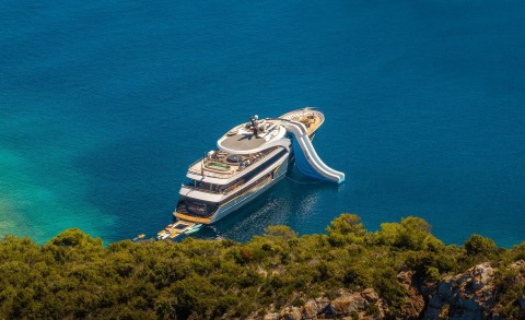 Luxury Bella yacht anchored in clear blue sea with a curved waterslide on deck near rocky shoreline