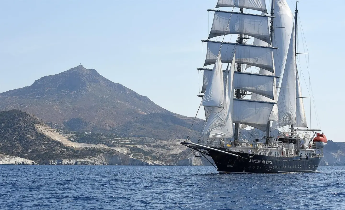 Yacht named Running on Waves sails near rugged hills at sunset on calm blue sea