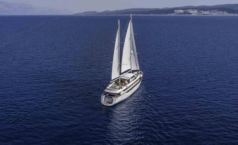 A white, two-masted sailing yacht sails in open blue sea with a distant coastline in the background