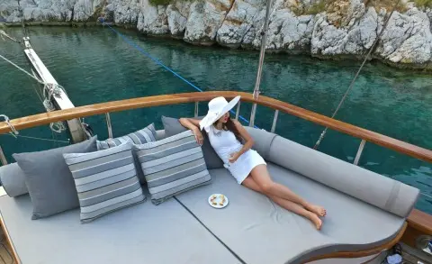 Person in white shirt and shorts lounging on aft deck of gulet Kaptan Mehmet Bugra near rocky coast