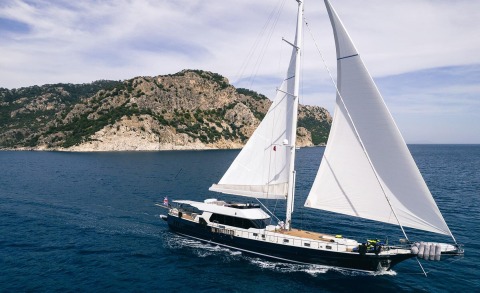 A large white-sail yacht on blue sea, with rocky coastline in the background