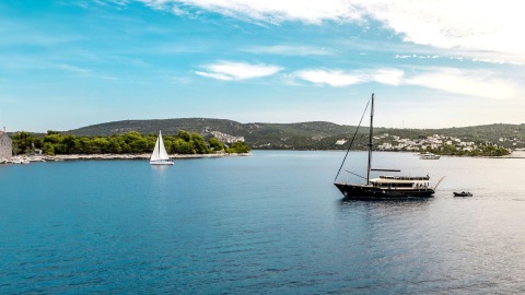 A dark sailboat anchored on blue water with green tree-lined shoreline