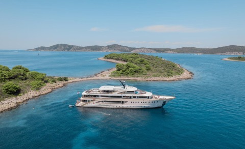 White Riva yacht anchored in clear blue sea near rocky islets, multiple decks visible