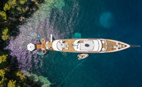 Aerial view of Maia yacht anchored by rocky coastline with crystal-blue water and a small tender nearby