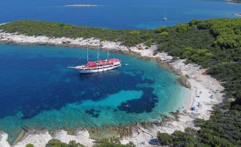Red and white three-masted yacht anchored by a rocky, tree-covered coastline