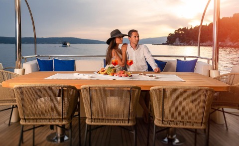 Couple with hat seated at a table on the deck, sunset view over the sea with platters of food