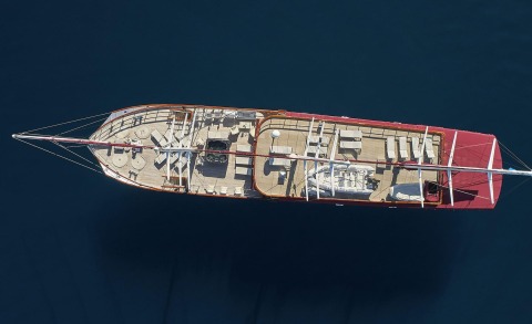Aerial view of Amorena yacht; teak deck with seating areas, tables and lounge spaces, red hull, and masts.