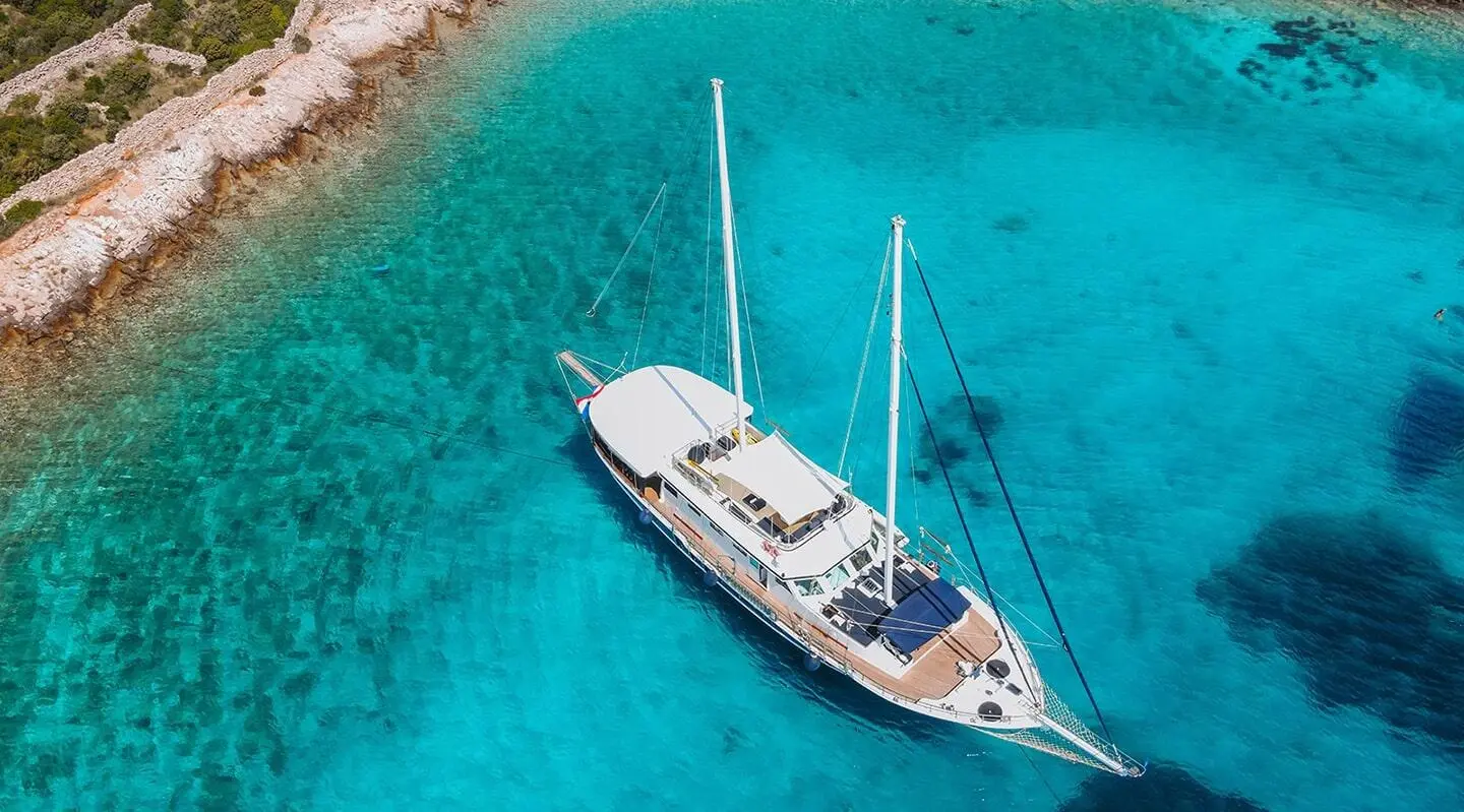 A white luxury sailing yacht Aurum anchored in clear blue water near rocky coastline