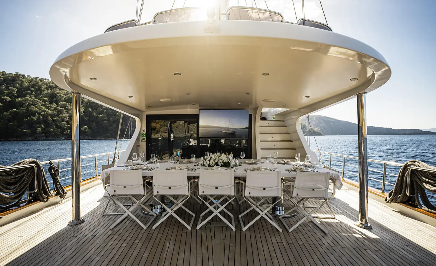Sunlit yacht rear deck with a long dining table, white chairs, and place settings