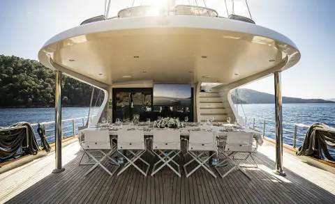 Sunlit yacht rear deck with a long dining table, white chairs, and place settings