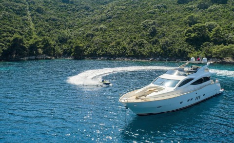 White luxury yacht anchored in turquoise sea near green hillside, small speedboat in wake
