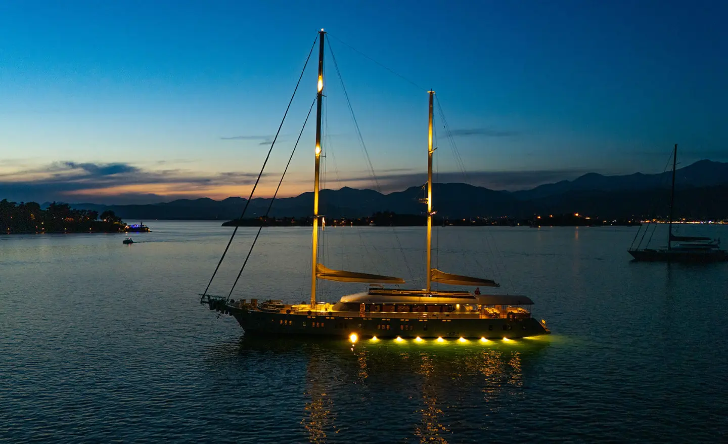 A motor yacht with two masts illuminated by warm yellow lights at night on the water