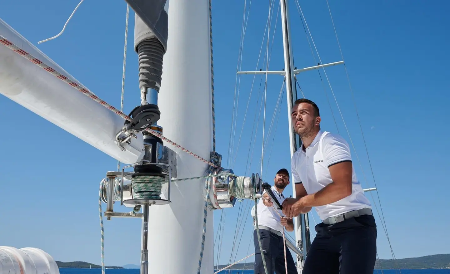 Two crew members on a sailing yacht near a mast with winches and ropes in close-up