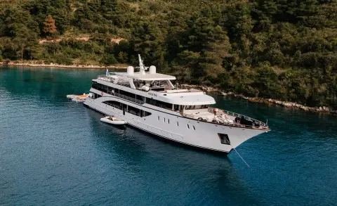 Cristal yacht anchored near forested coastline, white multi-deck vessel bobbing in blue water