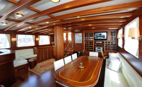 Interior of Kaya Guneri 4 yacht with large wooden table, white cushioned seating, and wood beams across ceiling, windows letting in natural light