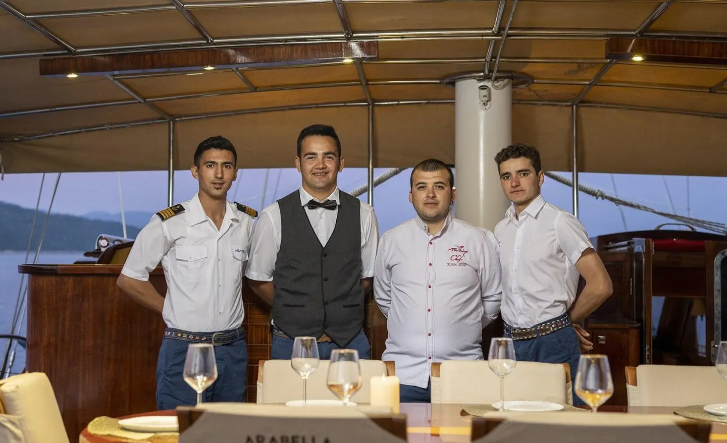 Crew members stand at a dining table on the Arabella gulet, with sea view behind