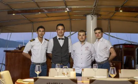 Crew members stand at a dining table on the Arabella gulet, with sea view behind
