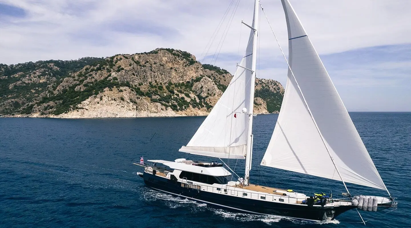 White luxury sailing yacht with large sails on blue sea near rocky coastline