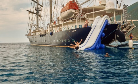 People jumping into the water beside a large inflatable slide attached to a large sailing yacht