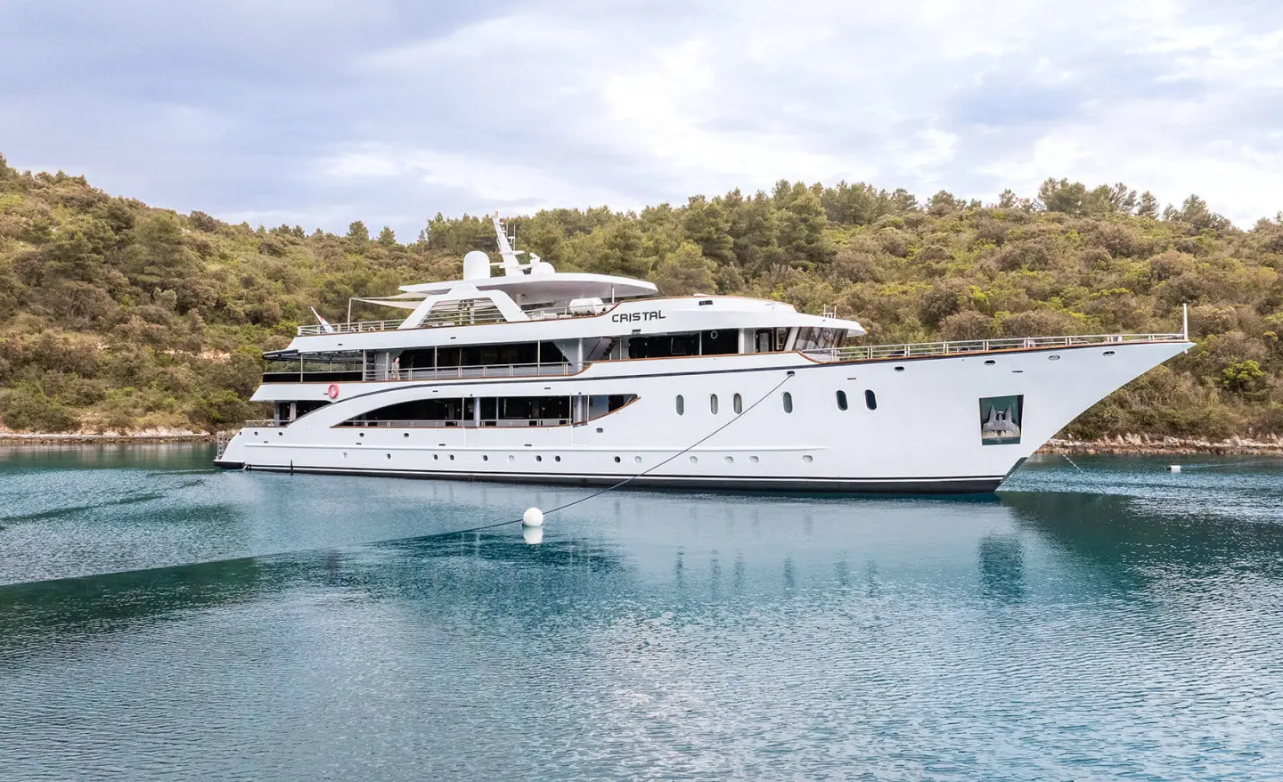 Motor yacht Cristal anchored near shore with white hull against green hill backdrop