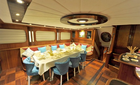 Berrak Su yacht interior dining area with wood paneling, plush blue chairs, table setting, and warm ambient lighting