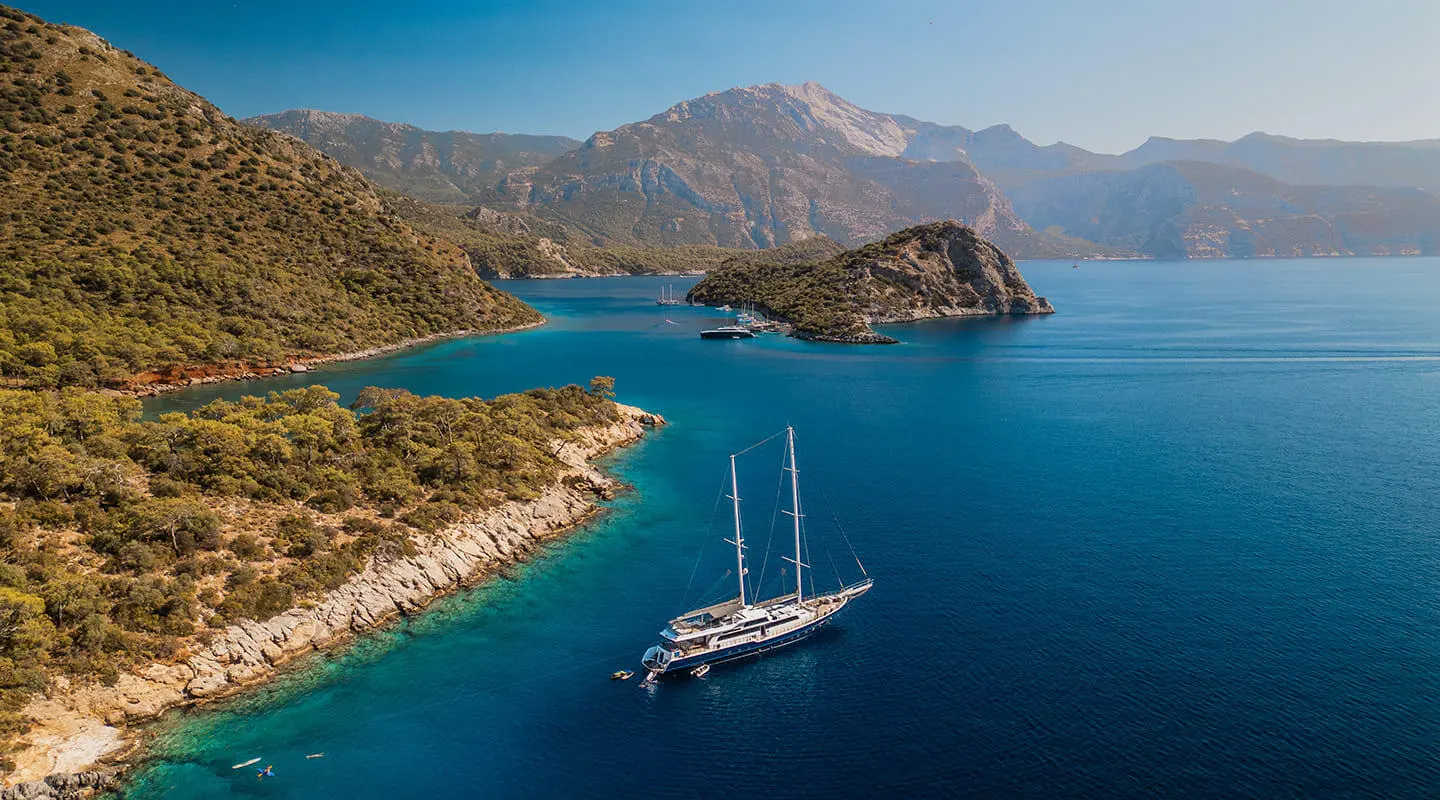 Esma Sultan yacht anchored in blue sea with a rugged coastline backdrop