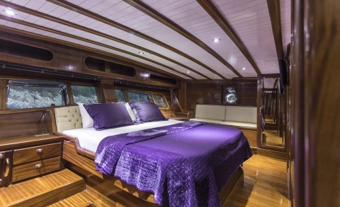 Grace 1 gulet with large bed, purple pillows and blanket in a wooden interior