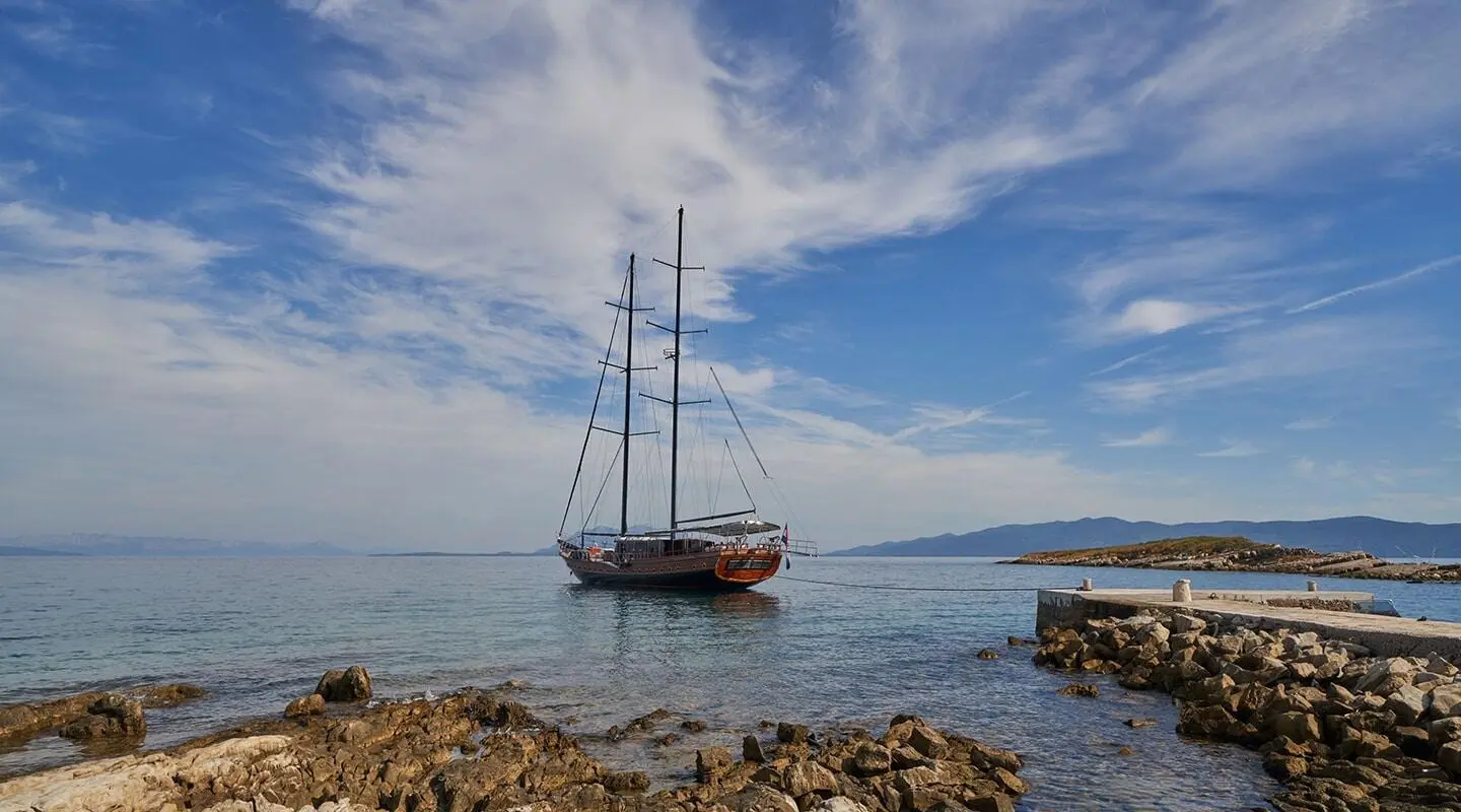 Sailing yacht anchored near rocky shore with a pier and calm sea.